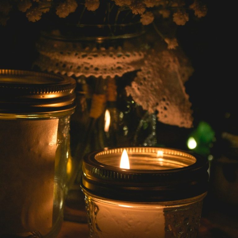 Faelorin - Wiccan Candles Candles glow softly among jars and decorative elements in a dimly lit setting.